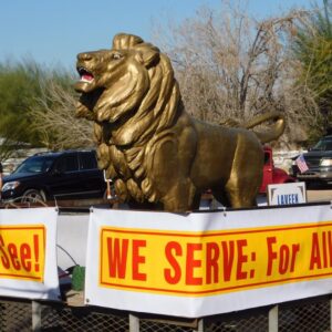 A stattue of a golden lion sitting on the bed of a float with the words WE SERVE wrapping around the truck bed.